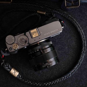 Op de afbeelding: Een zilveren Fujifilm X-Pro2 camera met een zwarte lens en een gevlochten zwarte leren camerariem. De riem heeft een goudkleurig accent met een "X" erop. De camera ligt op een donkergrijze ondergrond.