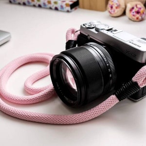 May include: A silver camera with a black lens and a pink camera strap. The camera has the text "X-E2" on the top.