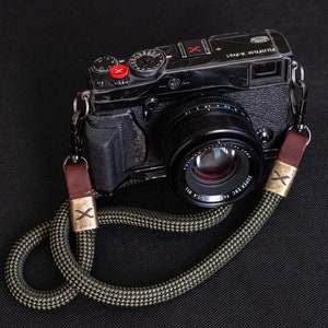 Könnte beinhalten: Eine schwarze Kamera mit einem schwarzen und grünen Kameragurt. Die Kamera hat ein Objektiv mit dem aufgedruckten Text "SUPER EBC FUJINON 1:1.4 35mm". Der Kameragurt hat eine Metallschnalle mit einem "X" darauf.