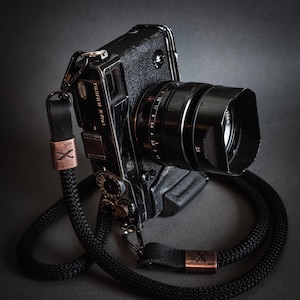 Puede incluir: Una cámara negra con una correa de cuero negra y una lente plateada. La cámara tiene una inscripción plateada que dice "FUJIFILM X-Pro1".