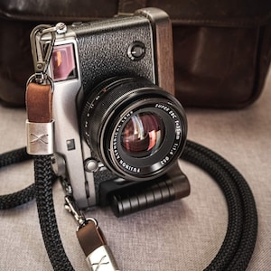Puede incluir: Una cámara plateada y negra con una empuñadura de madera marrón y una correa de cuero negra. El objetivo de la cámara es un Fujinon Super EBC 1:1.4 35mm.