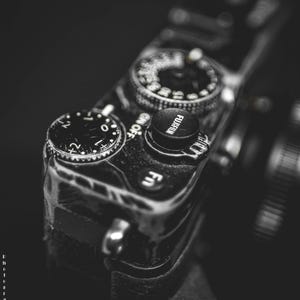 Puede incluir: Primer plano de una cámara en blanco y negro con varios diales y botones. La parte superior de la cámara presenta un botón con la marca "FUJIFILM". La imagen destaca los detalles de la cámara.