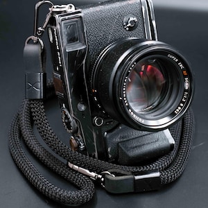 Może przedstawiać: Czarny aparat cyfrowy Fujifilm X-Pro1 z obiektywem 56 mm i czarnym, plecionym paskiem do aparatu. Obiektyw jest oznaczony jako "SUPER EBC XF 56mm 1:1.2 R 062". Pasek ma skórzany akcent i metalowe zapięcie.