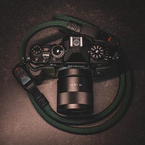 May include: A black Nikon Zf camera with a lens labelled FE 1.8/55 attached. The camera features a textured green grip and is connected to a thick, braided dark green and black rope camera strap with black leather connectors and red accents.