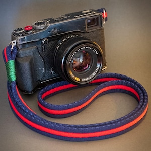 Può includere: Fotocamera nera con obiettivo e cinturino a righe blu, rosse e verdi. Il cinturino della fotocamera è ad anello e poggia su una superficie scura. L'obiettivo riporta la scritta "FUJINON ASPHERICAL LENS".