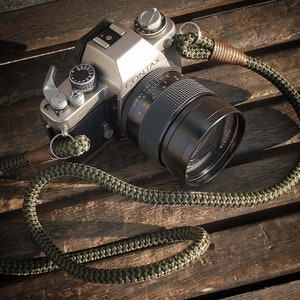 May include: A silver Contax camera with a black lens and a green and brown camera strap. The camera is sitting on a wooden surface.