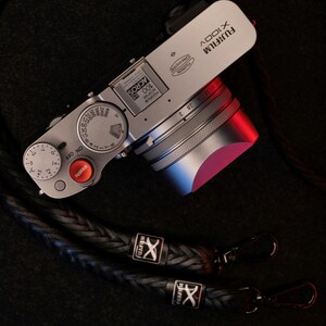 Può includere: Fotocamera digitale Fujifilm X100V argentata con cinturino in pelle intrecciata nera. La fotocamera ha un obiettivo argentato e un quadrante rosso e argento. Il cinturino ha chiusure in metallo e loghi Fujifilm.