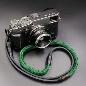 Puede incluir: Una cámara Fujifilm X-Pro2 plateada y negra con una lente negra y una correa verde y gris. La correa está hecha de un material verde tejido y cuero gris.
