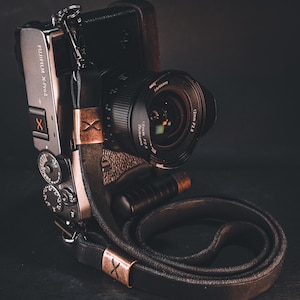 May include: A silver Fujifilm X-Pro2 camera with a black lens and leather strap. The lens is a 7Artisans 12mm f/2.8. The strap is black leather with copper-coloured accents. The camera is on a dark surface.