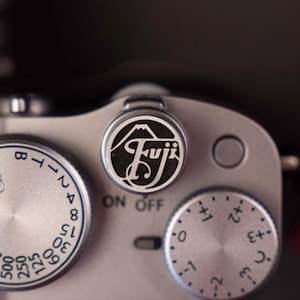 Puede incluir: Primer plano de un cuerpo de cámara plateado con diales y controles. El logotipo "Fuji" se muestra de forma destacada en un dial central, con el texto "ON OFF" debajo. Otros diales muestran marcas numéricas.