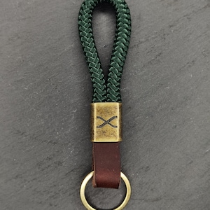 May include: A dark green rope keychain with a brass-colored metal accent featuring an "X" design. The keychain has a brown leather strap and a gold-colored ring.