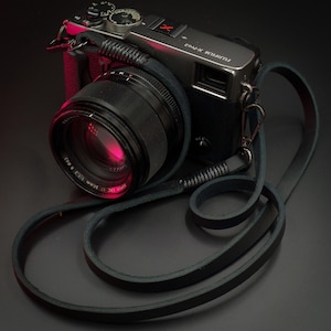 Może przedstawiać: Czarny aparat Fujifilm X-Pro2 z obiektywem 56mm f/1.2, wyposażony w długi czarny skórzany pasek ze wzmocnionymi szytymi końcami i metalowymi obrotowymi klipsami. Na korpusie aparatu widnieje napis Fujifilm X-Pro2 oraz czerwona ikona litery X.