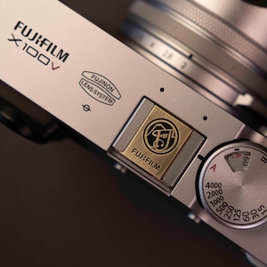 May include: Silver Fujifilm X100V digital camera with a black lens. The camera body features the Fujifilm logo, Fujinon Lens System text, and a dial with various settings. The camera is designed for photography.