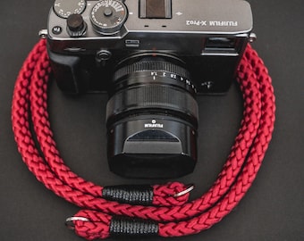 May include: A Fujifilm X-Pro2 camera with a black lens attached, positioned behind a coiled, braided red camera strap. The strap features black wrapped ends and silver metal rings for attachment, contrasting against the dark camera body.