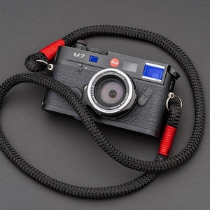 May include: A black Leica M7 film camera with a black lens and a red and black camera strap. The camera has a blue light meter and the text "M7" on the top.