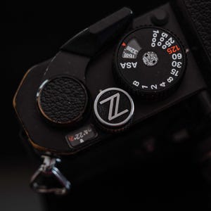 Puede incluir: Primer plano de una cámara negra con un dial negro y plateado y un botón plateado con un logotipo "Z". El dial muestra los ajustes ASA y las velocidades de obturación. También son visibles el agarre de cuero texturizado de la cámara y otros controles.