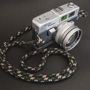 Könnte beinhalten: Eine silberne Petri Racer 35mm Filmkamera mit schwarzem Kunstledergehäuse und einem schwarz-grünen Kameragurt. Die Kamera hat ein silbernes Objektiv mit einer schwarzen Objektivkappe.