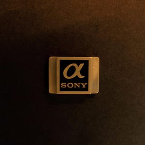May include: A gold-colored metal badge with the Sony logo and the Greek letter alpha. The badge is rectangular with rounded corners and is set against a dark brown background.