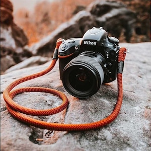 Puede incluir: Una cámara Nikon D800 negra con un objetivo, apoyada en una roca gris. La cámara está conectada a una correa de cámara naranja.