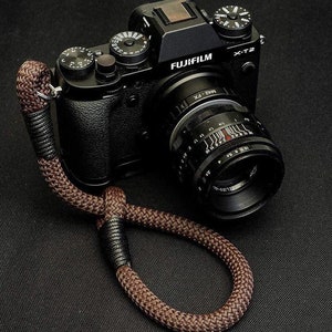 May include: A black Fujifilm X-T2 camera with a brown rope camera strap. The camera has a black lens with white markings and the text "FUJIFILM X-T2" on the top.