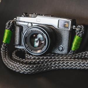 Pode incluir: Uma câmera prateada com uma lente preta e uma alça de câmera preta e verde. A lente diz "SUPER EBC FUJINON 35MM 1:1.4 ASPHERICAL LENS".