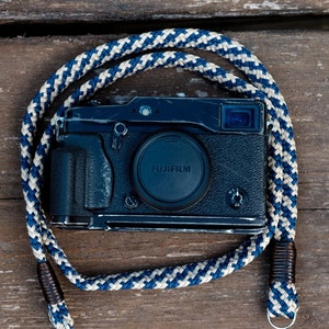 Puede incluir: Una cámara negra con una correa de cuerda azul y blanca. La cámara tiene un objetivo con la palabra "FUJIFILM" impresa en él.