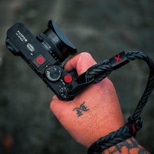 Könnte beinhalten: Eine schwarze Fujifilm X100V Digitalkamera mit einem schwarzen geflochtenen Leder-Handschlaufe. Die Kamera hat rote Akzente am Auslöser und am oberen Einstellrad, wobei der Text Fujifilm X100V sichtbar ist. Die Schlaufe enthält einen schwarzen Metallverbinder mit einem roten X.