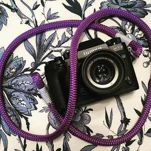 Può includere: Una macchina fotografica nera con una tracolla viola. La macchina fotografica ha un obiettivo con il testo "FUJIFILM X-T2 FUJINON ASPHERICAL LENS" su di esso.