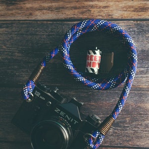 Può includere: Una macchina fotografica Zenit nera con una tracolla in corda blu e bianca. La macchina fotografica ha un'etichetta 12xp e un rullo di pellicola con un'etichetta "Street Candy" è accanto.