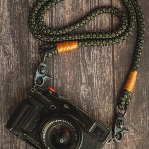 May include: Black camera with a green rope strap and orange accents. The camera has a lens with the text "FUJINON ASPHERICAL LENS" and "SUPER EBC". The strap is attached to the camera with metal clips. The background is a dark wood surface.