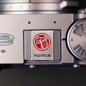 Puede incluir: Primer plano de una cámara digital Fujifilm plateada. La cámara presenta un logotipo rojo y blanco con el nombre de la marca "FUJIFILM" debajo. Un dial con números y ajustes es visible en el lado derecho de la cámara.