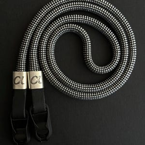 May include: A black and white patterned camera strap. The strap is made of a thick, woven material and features black leather connectors and silver metal accents with the Greek letter alpha. The strap is designed to be looped around the neck.