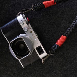 Op de afbeelding: Een zilveren camera met een zwarte leren band en rode accenten. De camera heeft een lensdop en een zoeker.
