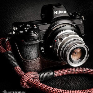 Op de afbeelding: Een zwarte Nikon camera met een zilveren lens bevestigd. De lens is gemarkeerd met "Ernst Leitz GMON Elmar F-9cm 1:4". De camera is bevestigd aan een rood en zwart touw.