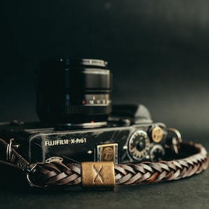 Peut inclure: Un appareil photo Fujifilm X-Pro1 noir avec un objectif et une sangle de caméra tressée en cuir marron. La sangle a un fermoir en métal doré et une pièce métallique rectangulaire avec un "X". L'appareil photo est sur une surface sombre.
