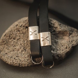 Könnte beinhalten: Nahaufnahme von zwei schwarzen Leder-Schlüsselanhängern mit silberfarbenen Metallakzenten. Jeder Schlüsselanhänger hat ein rechteckiges Metallstück mit einem "X"-Design. Die Schlüsselanhänger sind an silberfarbenen Schlüsselringen befestigt.