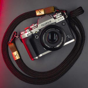 May include: A silver and black Fujifilm X-T3 camera with a Fujinon Aspherical Lens Super EBC 35mm f/1.4 lens attached. The camera features a red shutter button and is connected to a thick, black braided rope camera strap with brown leather accents.