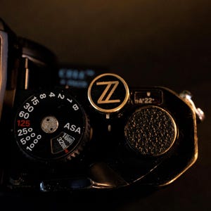Puede incluir: Primer plano de una cámara vintage negra con un logotipo 'Z' dorado. La cámara presenta un dial con números y ajustes ASA, y un botón texturizado. El fondo es oscuro.