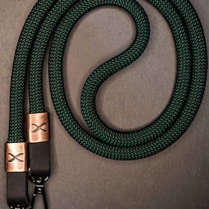 Op de afbeelding: Een lang, gevlochten touwkoord met een donkergroen en zwart geweven patroon. De uiteinden zijn vastgezet met koperkleurige metalen hulzen, elk voorzien van een X-symbool, en bevestigd aan zwarte leren lipjes met zwarte metalen draaibare haken.