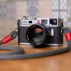 May include: A silver Leica M9 camera with a black lens and a red button on the top. The camera is attached to a gray and red camera strap.