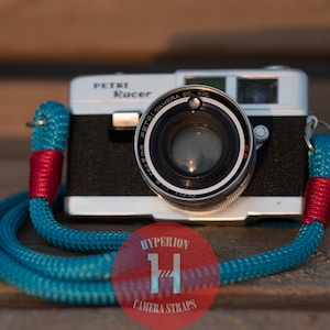 Può includere: Una macchina fotografica a pellicola da 35 mm di colore argento e nero con una tracolla blu e rossa. La macchina fotografica ha un obiettivo con il testo "PETRI CAMERA CO. INC." e "PETRI Racer" stampato sul davanti. La tracolla è una corda blu con accenti rossi.