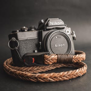Op de afbeelding: Een zwarte Nikon camera met een bruine, gevlochten leren polsriem. De camera heeft een lensdop met het Nikon logo. De riem is om de camera gewikkeld. De achtergrond is donker.
