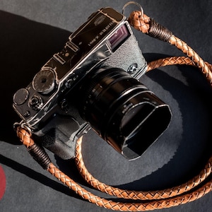 May include: A silver camera with a black lens and a brown leather camera strap. The strap is braided and looped around the camera. The camera is on a dark surface. A red circle with white text that says "Hyperion Camera Straps" is in the lower left corner.