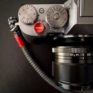 Puede incluir: Primer plano de una cámara Fujifilm plateada con detalles en negro. La cámara tiene múltiples diales y un botón rojo con el logotipo de Fujifilm. La lente es negra y la correa de la cámara es negra y roja.