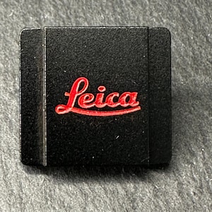 May include: A black rectangular camera accessory with the red Leica logo. The logo is in a cursive font. The accessory is on a grey, textured surface.