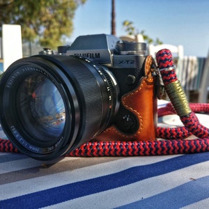 Könnte beinhalten: Eine silberne Fujifilm X-T2 Kamera mit einem schwarzen Fujinon 56mm f/1.2 R Objektiv. Die Kamera befindet sich in einem braunen Lederetui und hat eine rote und blaue Seilkamera-Schlaufe.