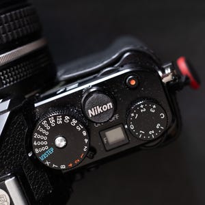 Puede incluir: Primer plano de una cámara Nikon negra con varios diales y botones. La cámara cuenta con una lente, un dial de velocidad de obturación con números y un dial superior con el logotipo de Nikon. Un botón rojo es visible en el lateral.