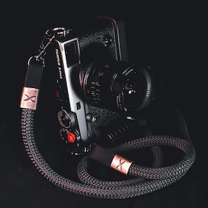 Może przedstawiać: Srebrny aparat Fujifilm X-Pro2 z czarnym obiektywem i szaro-czarnym paskiem do aparatu z liny. Pasek ma miedziane akcenty i czarny skórzany łącznik. Aparat ma czerwony przycisk i napis "FUJIFILM X-Pro2".