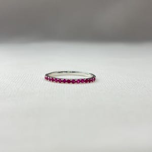 May include: A thin silver band ring featuring a continuous row of small, round, vibrant pink gemstones set along the outer circumference. The metal has a polished, reflective finish, and the stones are uniform in size and color throughout the band.
