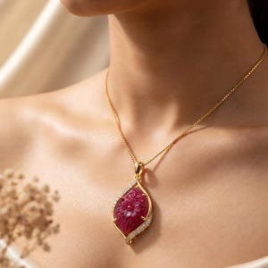Puede incluir: Un collar de cadena dorada con un colgante en forma de marquesa que presenta una piedra preciosa de color rojo intenso tallada con motivos florales. El colgante está enmarcado por pequeñas piedras claras y brillantes.